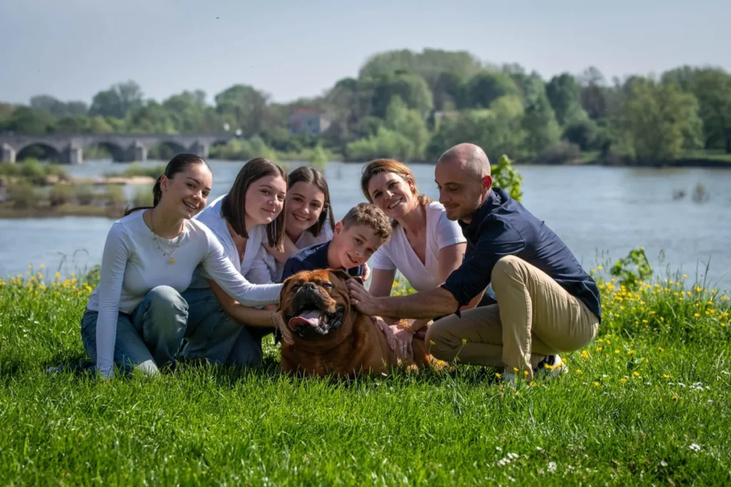 Famille souriante (6 personnes) avec leur grand chien marron, caressant l'animal sur l'herbe verte au bord d'une rivière.