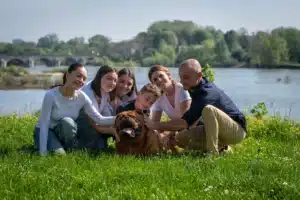 Famille souriante (6 personnes) avec leur grand chien marron, caressant l'animal sur l'herbe verte au bord d'une rivière.
