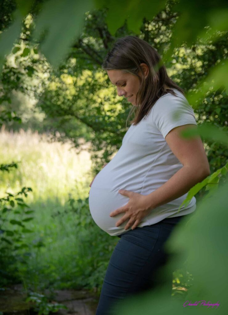 Femme enceinte souriante en t-shirt blanc, tenant tendrement son ventre rond, entourée d'une nature verdoyante. Chantal Photography.